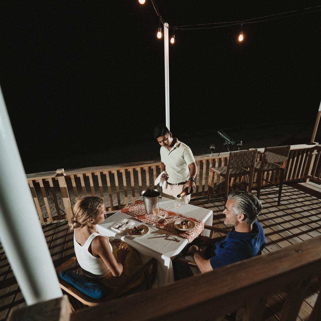 A waiter pours champagne for a man and a woman enjoying dinner on a terrace under the night sky.
